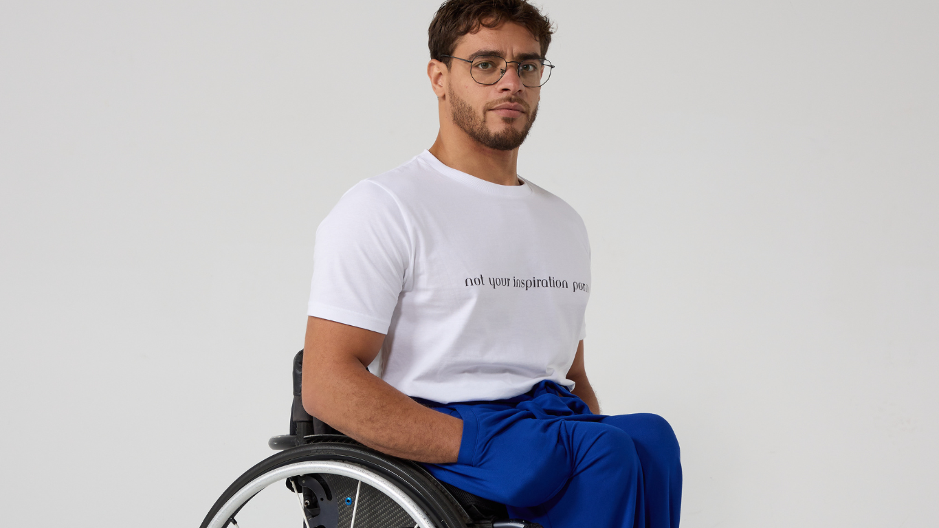 a man with a beard and round glasses is modelling a white tshirt and blue trousers in his manual wheelchair. He has his hands in his pockets a man with a beard and round glasses is modelling a white tshirt and blue trousers in his manual wheelchair. He has his hands in his pockets
