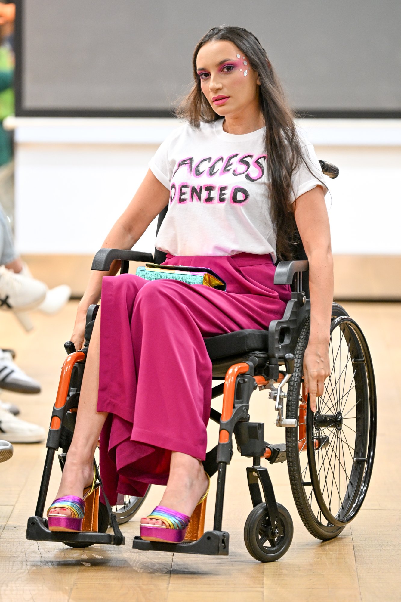 a woman with long brown hair is posing on a catwalk in her orange and black manual wheelchair. She is wearing a white tshirt saying "access denies", pink trousers and rainbow heeled shoes a woman with long brown hair is posing on a catwalk in her orange and black manual wheelchair. She is wearing a white tshirt saying "access denies", pink trousers and rainbow heeled shoes