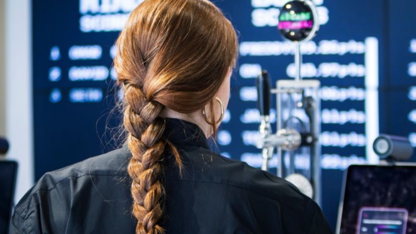 the back of a woman's head. She has a plait in her long dark hair and is looking at a beer pouring robot and large scoreboard screen in front of her